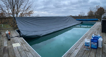 A backyard swimming pool with a winter cover being installed across the water, surrounded by pool-closing tools on the deck under an overcast autumn sky.