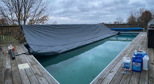 A backyard swimming pool with a winter cover being installed across the water, surrounded by pool-closing tools on the deck under an overcast autumn sky.